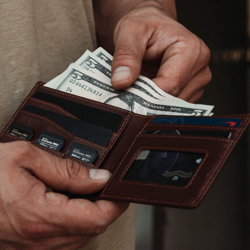A person holds an open brown wallet containing cash, several credit cards, and SD memory cards, with one hand holding the wallet and the other hand placing or removing a five-dollar bill.