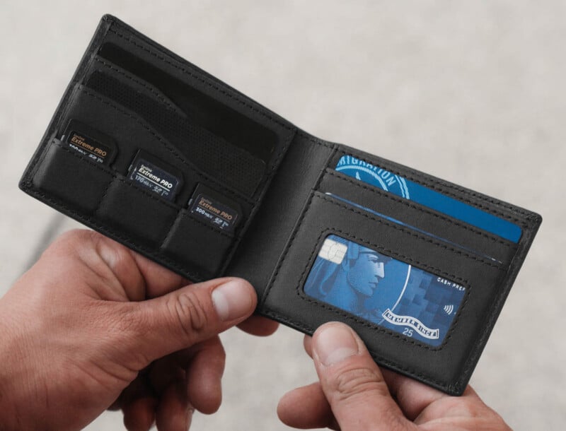 A person holds an open black wallet containing memory cards, a visible blue credit card, and another card with a partially exposed chip. The wallet is held with both hands against a blurred background.