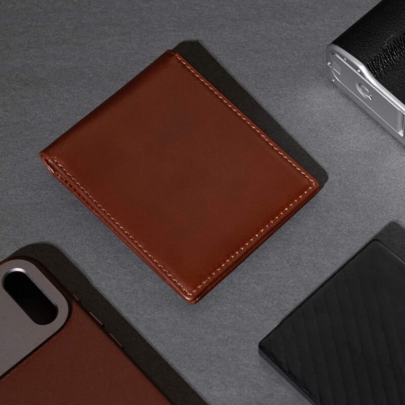 A brown leather wallet lies on a gray surface, surrounded by a smartphone with a brown case, a black textured object, and a metallic device in the corner.