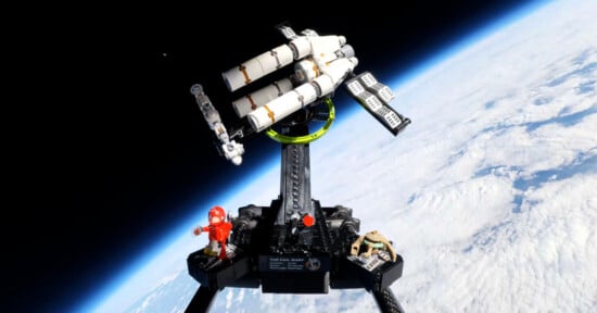 A LEGO space shuttle model with astronaut and alien figures is positioned on a stand, photographed against the backdrop of Earth’s curved horizon and clouds from high altitude.