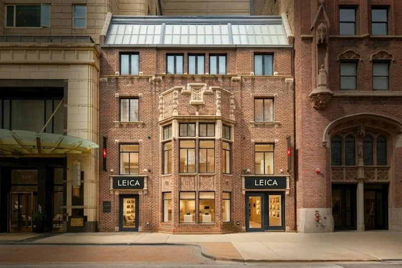 A three-story brick building with large windows and two doors, displaying signs that read "Leica." The building is flanked by taller historic structures on either side on a city street.