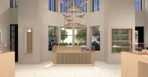 Modern retail store interior with a light wood counter in the center, minimalist shelves, large windows letting in natural light, and a circular, layered chandelier hanging from the ceiling. Trees are visible outside.