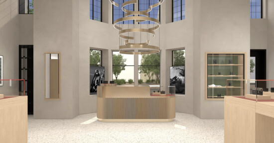 Modern retail store interior with a light wood counter in the center, minimalist shelves, large windows letting in natural light, and a circular, layered chandelier hanging from the ceiling. Trees are visible outside.