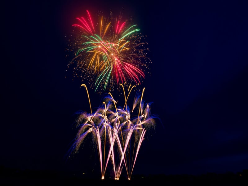 Colorful fireworks burst in the night sky, with red, green, yellow, and blue sparks radiating above a dark landscape, creating vibrant trails and bright patterns against a deep blue background.