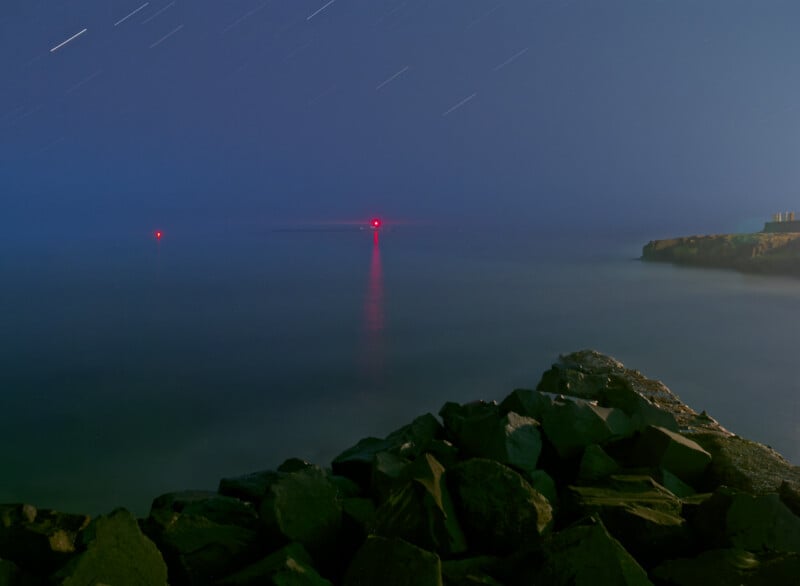 A rocky shoreline at night under a starry sky, with two red lights in the distance reflecting on calm water; faint star trails are visible, and a cliff is seen on the right side of the image.