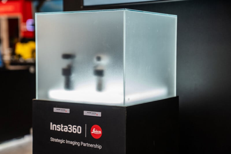 A display case with frosted glass showcases two blurred objects inside. The label on the base reads "Insta360" and "Leica Strategic Imaging Partnership." The background is out of focus.
