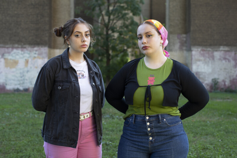 Two young women stand outdoors on grass in front of a worn, graffiti-covered wall. One wears a black jacket and pink pants; the other wears a colorful scarf, green top, and blue jeans. Both look seriously at the camera.