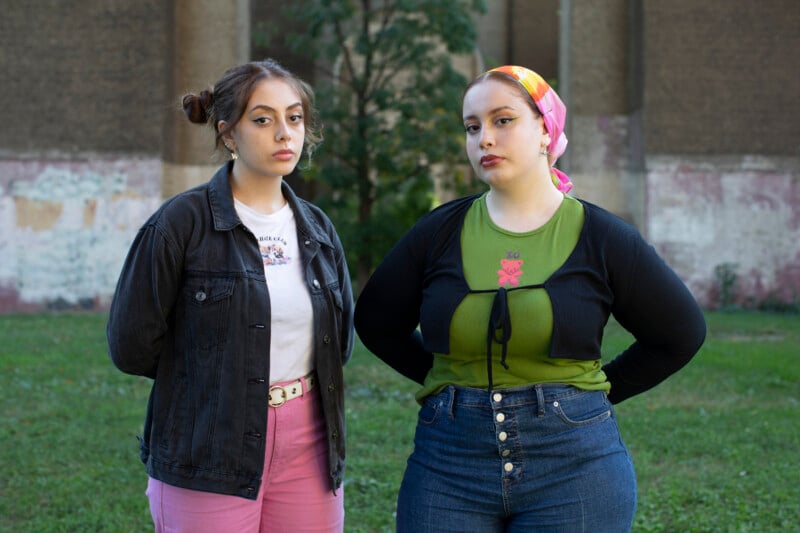 Two young women stand outdoors on grass in front of a worn, graffiti-covered wall. One wears a black jacket and pink pants; the other wears a colorful scarf, green top, and blue jeans. Both look seriously at the camera.