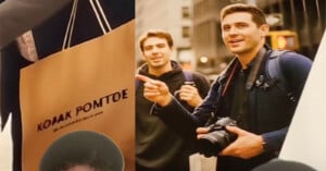 Two men stand outdoors in a city; one holds a camera and points, while the other smiles. A close-up of a tan shopping bag with distorted “KODAK ROMTOE” text appears on the left side of the image.