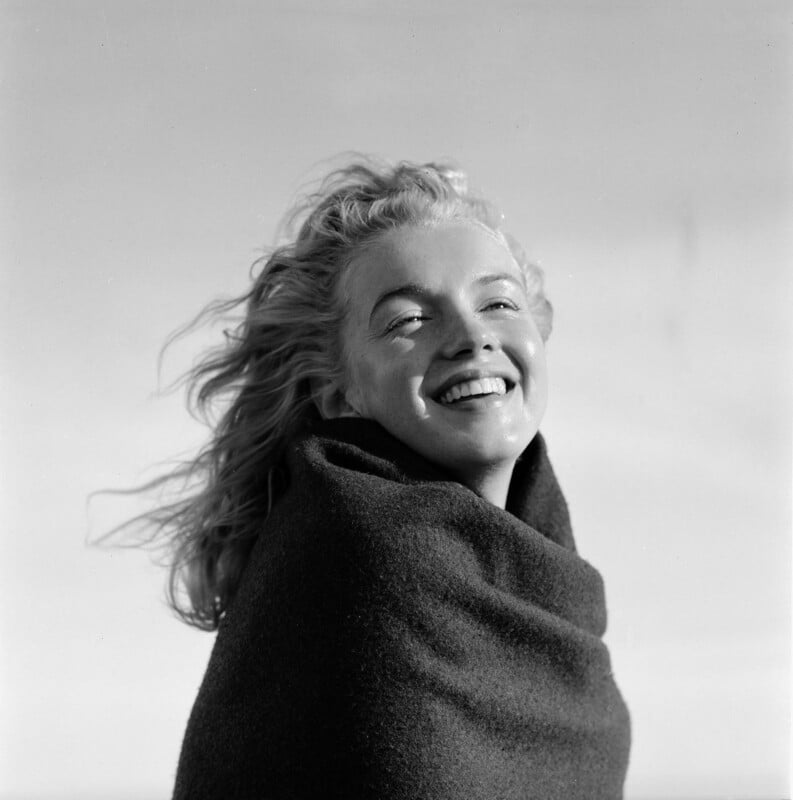 A woman with wavy, windblown hair smiles brightly at the camera, wrapped in a dark blanket against a light, blurred background, creating a joyful and carefree mood.