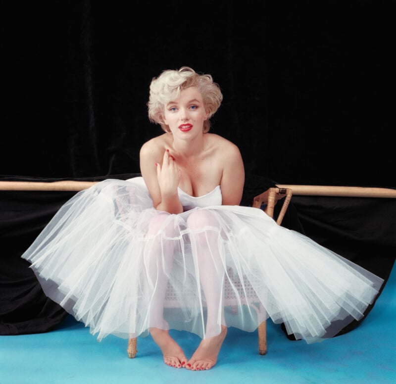 A woman with short blonde hair and red lipstick sits barefoot on a chair, wearing a strapless white tulle dress. She poses against a black backdrop and blue floor, gazing directly at the camera with a soft expression.