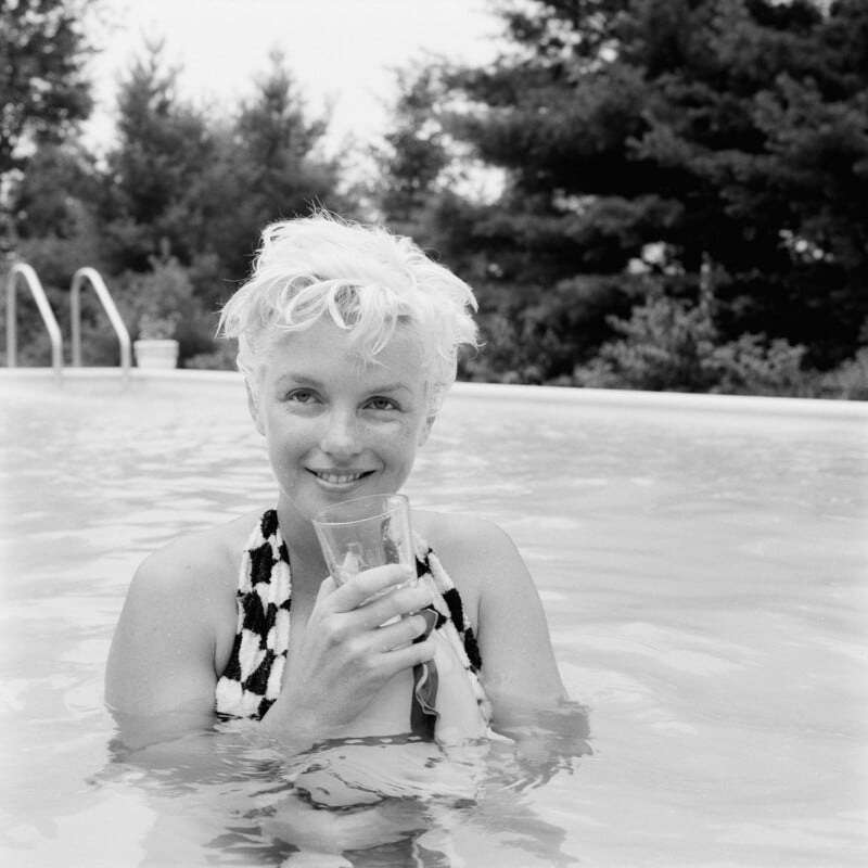 A woman with short, light-blond hair stands in a swimming pool, smiling and holding a glass. She wears a patterned towel or garment draped around her shoulders, with trees visible in the background.