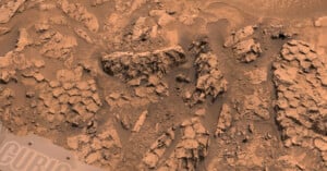 A close-up view of a rocky, reddish Martian surface with jagged, layered rock formations and scattered dust. Part of a rover’s equipment marked “CURI” is visible in the bottom left corner.