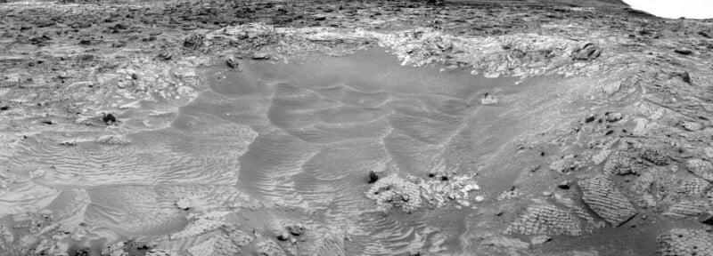 Black and white image of a shallow, sandy crater on a rocky, barren landscape, with rippled sand patterns and scattered rocks, resembling the surface of Mars.