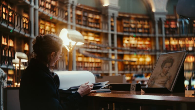 A person with glasses sits at a desk in a grand library, reading a book. Shelves filled with books curve around the well-lit, spacious room. A lamp and an illustrated portrait are visible on the desk.