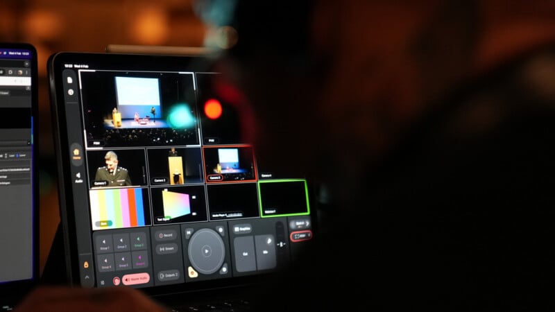 A close-up of a person operating a video switcher interface on a touchscreen device, displaying multiple live video feeds from an event with a stage and speaker.