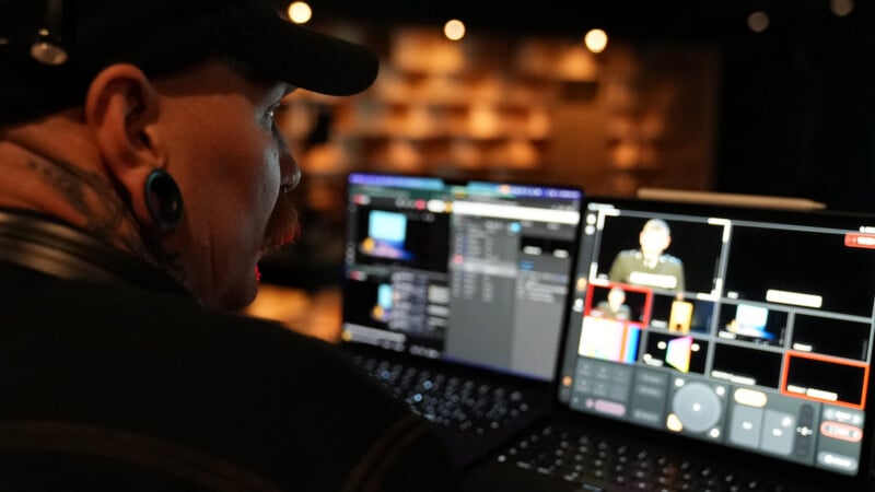 A person with tattoos and piercings monitors a video production setup, looking at multiple screens displaying video feeds and editing controls in a dimly lit room.