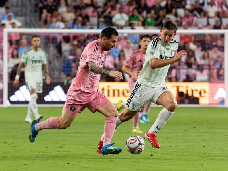 Two soccer players chase the ball during a match, one in a pink kit and the other in a white and green kit, with a blurred crowd and other players in the background on a grassy field.
