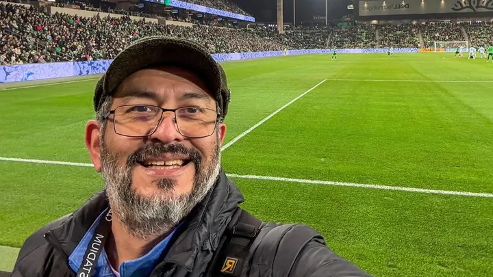A man wearing glasses, a cap, and a jacket smiles for a selfie at a soccer stadium, with the green field and packed stands full of spectators in the background.