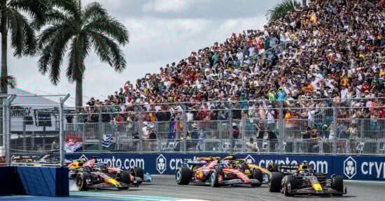 Formula 1 cars race around a track corner as a large crowd of spectators watch from the grandstands under palm trees on a sunny day. Advertising banners line the fence alongside the track.