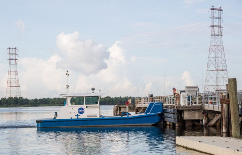Un barco azul y blanco de la NASA estaba atracado en el muelle en aguas tranquilas, y un hombre estaba parado en el muelle. Al fondo se ven dos altas torres de energía bajo un cielo parcialmente nublado.