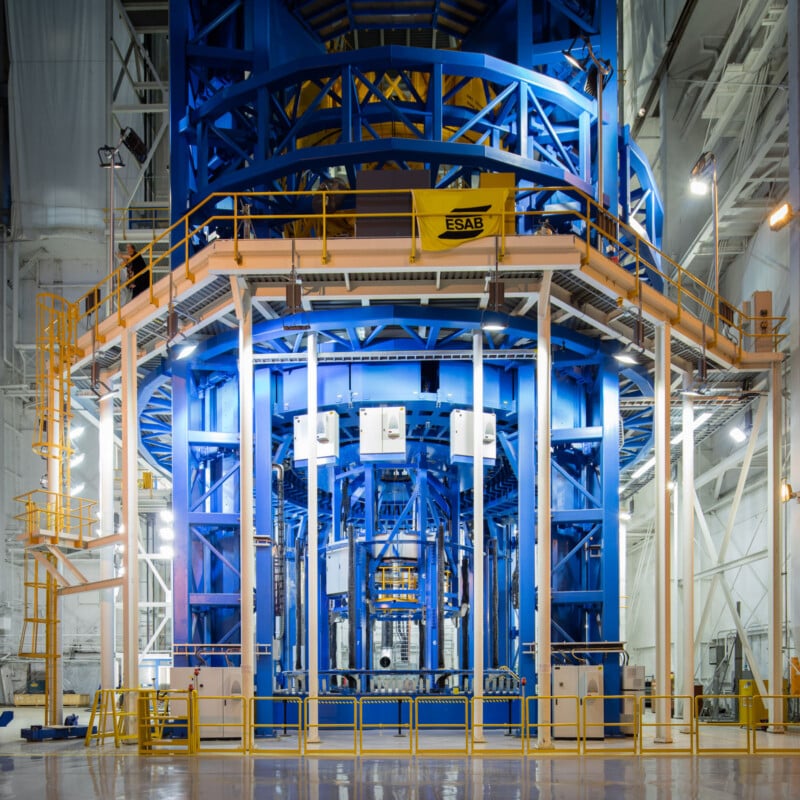 Una gran estructura industrial de metal azul se encuentra dentro de las instalaciones brillantemente iluminadas, rodeada de barandillas, plataformas y escaleras amarillas, con varias tuberías y equipos claramente visibles.