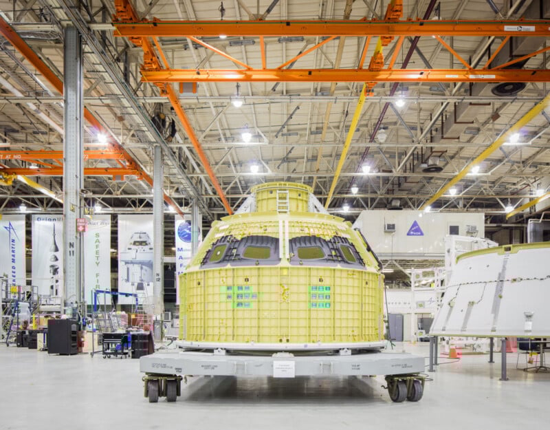 El módulo de la nave espacial de aspecto amarillo se exhibe en una plataforma en una instalación industrial espaciosa y luminosa, con techos altos, vigas de metal, maquinaria y el logotipo de la NASA claramente visible al fondo.
