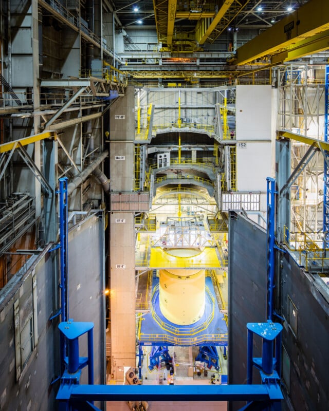 Interior de una gran instalación industrial con plataformas, pasillos y barandillas de metal amarillo rodeadas de piezas de máquinas cilíndricas donde los trabajadores ensamblan varios pisos y equipos en un área espaciosa y de techos altos.