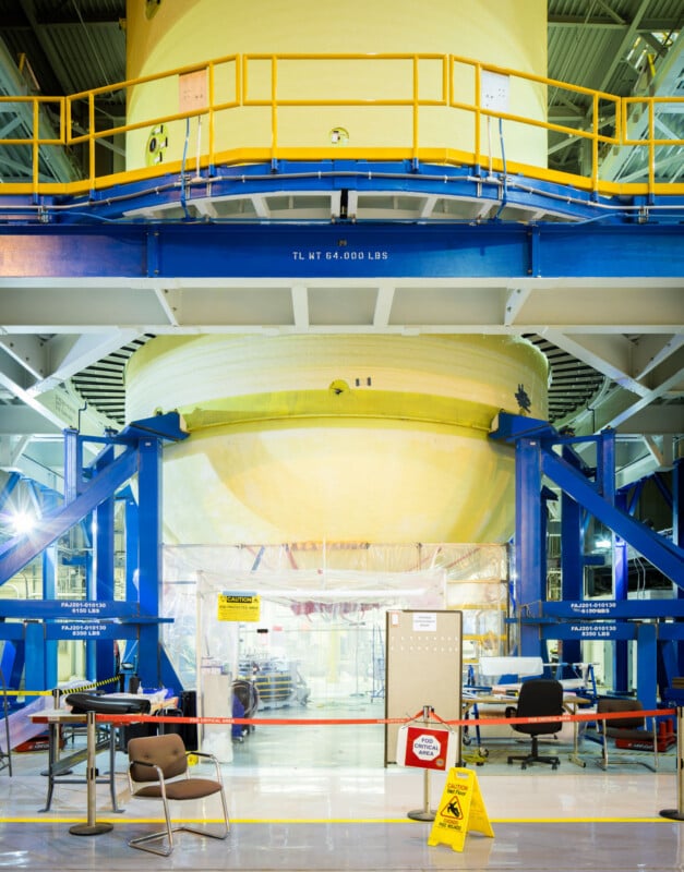 Un gran componente de cohete cilíndrico de color amarillo cuelga sobre el suelo dentro de una instalación industrial y está sostenido por estructuras azules. En primer plano se ven sillas, mesas y carteles de advertencia.