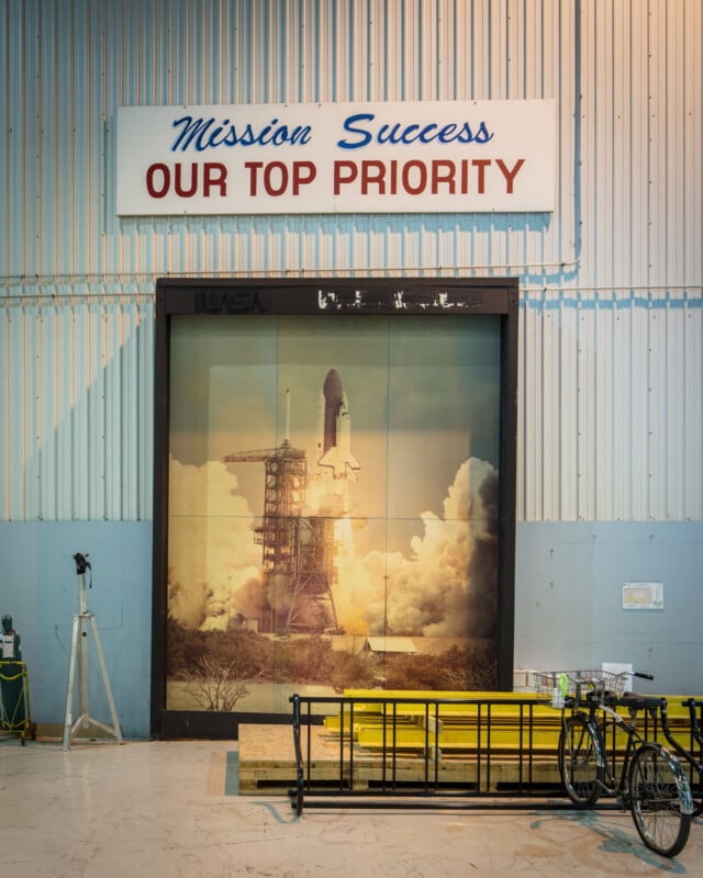 A large sign reads "Mission Success OUR TOP PRIORITY" above a wall mural of a space shuttle launching. In front, there are benches, a bicycle, and some equipment in an industrial setting.