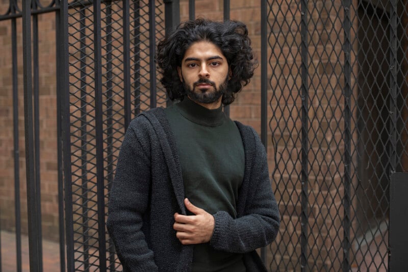 A man with curly hair and a beard stands in front of a metal fence, wearing a dark green shirt and a textured dark cardigan. He looks calmly at the camera, with one hand holding his cardigan. A brick wall is in the background.