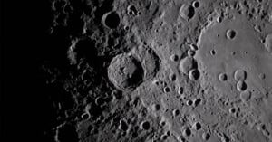 A detailed, close-up view of the moon’s surface shows numerous craters of varying sizes, with shadows accentuating their depth and texture. The image is in grayscale, highlighting the rugged lunar landscape.