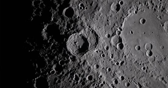 A detailed, close-up view of the moon’s surface shows numerous craters of varying sizes, with shadows accentuating their depth and texture. The image is in grayscale, highlighting the rugged lunar landscape.