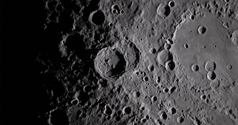 A detailed, close-up view of the moon’s surface shows numerous craters of varying sizes, with shadows accentuating their depth and texture. The image is in grayscale, highlighting the rugged lunar landscape.