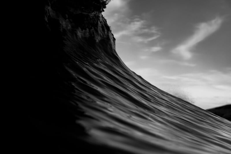 A black and white photo of a large ocean wave, captured from a low angle, with rippling water in the foreground and a cloudy sky in the background.