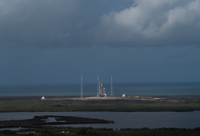 A distant view of a rocket standing upright on a launch pad near the coastline, with a cloudy sky above, water in the foreground, and the ocean in the background.