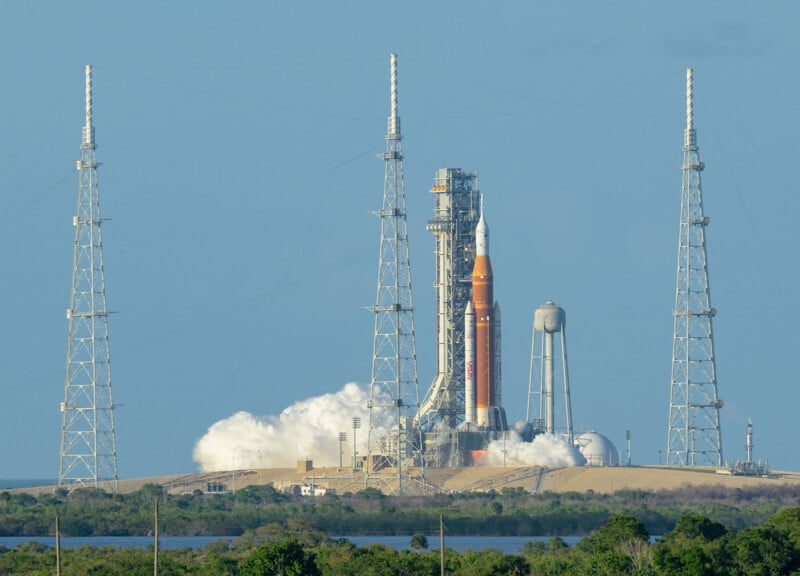 A large rocket on a launch pad is surrounded by four tall support towers. White smoke billows from its base, with a blue sky and some greenery in the foreground.