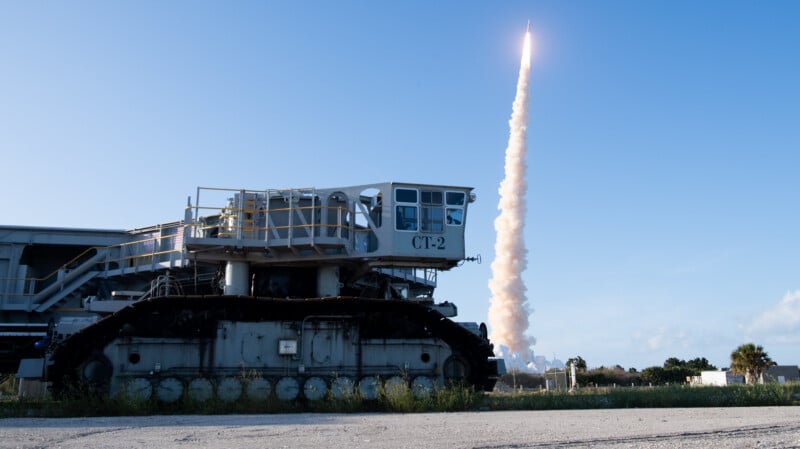 A large, tracked NASA crawler-transporter labeled "CT-2" sits on grass, while a rocket launches in the background, leaving a trail of smoke against a clear blue sky.