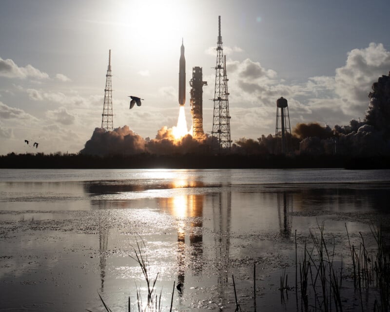 A rocket launches into the sky at sunrise, surrounded by large towers and billowing smoke, with birds flying and its reflection visible in a calm body of water in the foreground.