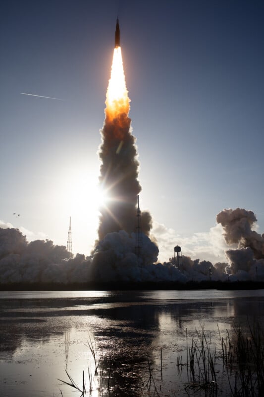 A rocket launches into the sky, trailing bright flames and smoke, silhouetted against the sun. Reflections and tall grasses are visible in the water in the foreground, with towers and clouds in the background.