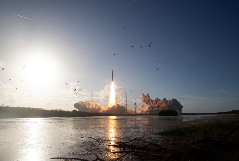 A rocket launches into the sky, leaving a trail of smoke and fire, with birds flying overhead and the sun shining brightly over a reflective body of water in the foreground.