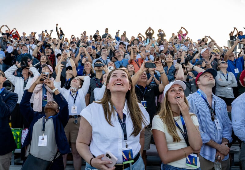 A large crowd of people stand and sit closely together, looking up and smiling, with many holding up phones and cameras to capture something in the sky during the daytime.