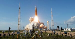 A large rocket launches into the sky, leaving a trail of smoke and fire. The launchpad is surrounded by cameras and equipment, with blue sky and light clouds in the background.