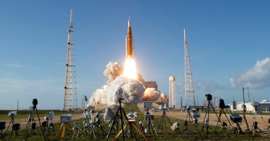 A large rocket launches into the sky, leaving a trail of smoke and fire. The launchpad is surrounded by cameras and equipment, with blue sky and light clouds in the background.