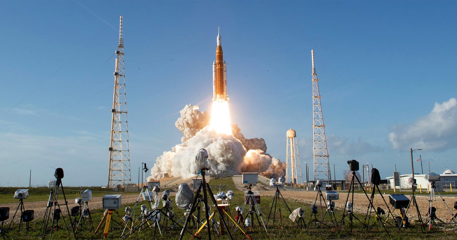 A large rocket launches into the sky, leaving a trail of smoke and fire. The launchpad is surrounded by cameras and equipment, with blue sky and light clouds in the background.