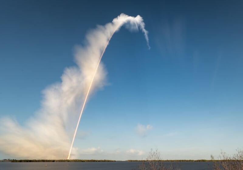 A rocket launches near the coast, leaving a bright, curved trail through a blue sky with streaks of clouds drifting upwards and outward from the launch site. Water and land are visible in the foreground.