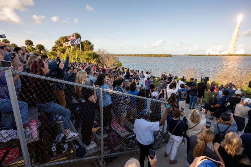 A large crowd gathers on bleachers by a waterfront, watching and photographing a rocket launch as a trail of smoke rises into the sky in the distance. The scene is lively under a clear, sunny sky with scattered clouds.