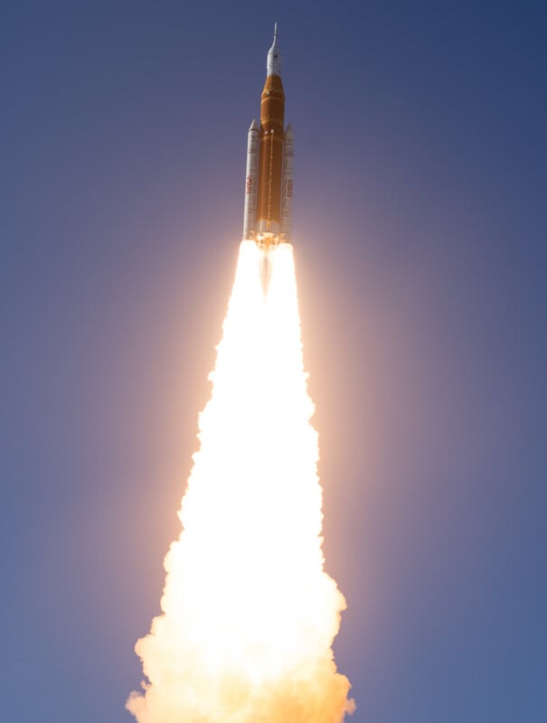 A rocket launches into a clear blue sky, emitting bright flames and smoke from its engines as it ascends vertically.