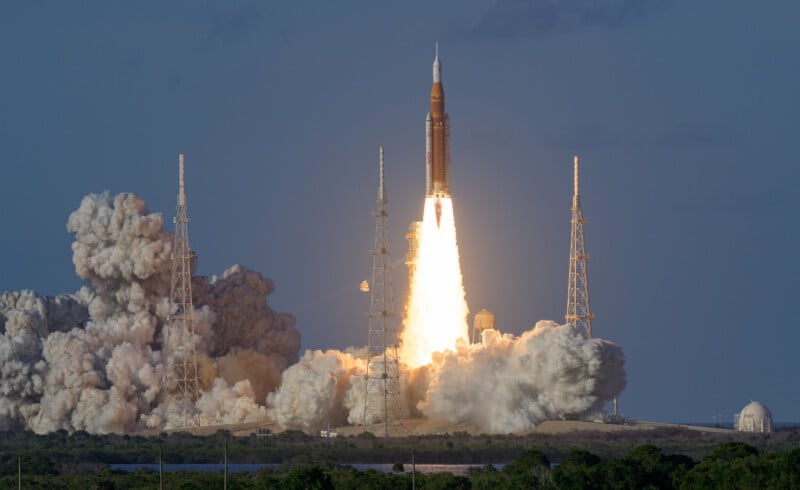 A rocket lifts off from a launch pad, emitting bright flames and thick smoke, surrounded by support towers against a clear sky.