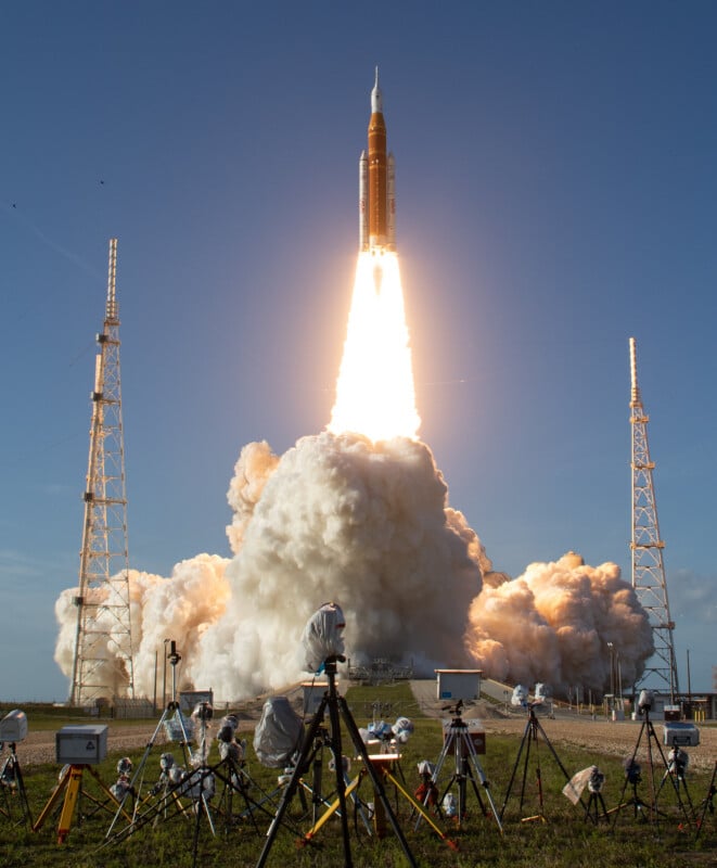 A rocket launches into the sky, emitting bright flames and smoke, while surrounded by support structures and cameras on tripods capturing the event. The sky is clear and blue.
